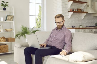 Genç, beyaz bir adam evdeki kanepede oturup dizüstü bilgisayarla not alıyor. Erkek çalışan işadamı internette bilgisayar danışmanlığı yapıyor. Uzak meslek, teknoloji, serbest meslek kavramı.