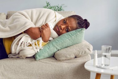 Grip belirtileri gösteren hasta bir genç kadın. Kareli yastığa sarılı, rahat bir odada yatan çekici Afrikalı Amerikalı kız. Soğuk ve mevsimsel hastalıklar