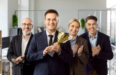 İş adamı lideri, iş dünyası kutlamaları sırasında kupayı elinde tutuyor. Gülümseyen iş kadını ve meslektaşları, parlak bir ofiste ödülü kazananı alkışlayarak başarının sinyallerini verdiler. Takım liderliği başarı konsepti.
