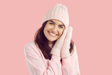 Gülümseyen mutlu esmer kadın rahat örgü şapkalı ve eldivenli pembe arka planda pembe sweatshirt 'lü kameraya bakıyor. Kış sonbahar tarzı, reklam afişi, insani duygular konsepti.