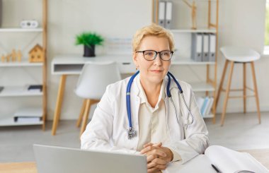 Son sınıfta gülümseyen, gözlüklü, beyaz üniformalı ciddi bir kadın doktor bilgisayarın yanında oturmuş, klinikteki hastaya uzaktan profesyonel danışmanlık yapıyor.