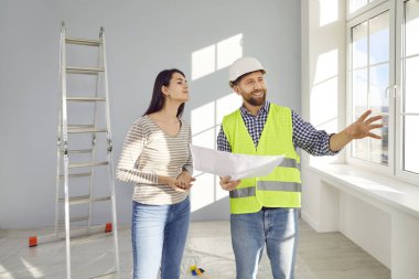 Erkek müteahhit bir kadınla konuşuyor. Yeni evde beyaz plastik pencereli, elinde inşaat kâğıdı planı olan ve genç ev sahibiyle konuşan kasketli ve üniformalı mutlu bir ustabaşı.