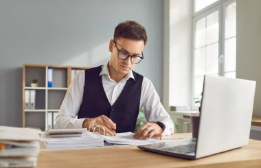 Harcamaları hesaplamak için dizüstü bilgisayar ekranına bakan ciddi bir adam, genç muhasebeci, faturaları internet üzerinden ödeyen, şirket bütçesini yöneten, mali evrak işleriyle meşgul biri.