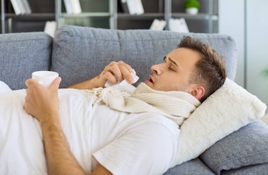 Ateşli hasta adam kanepede yatıyor. Evde, mendil ve bir kupa çay kullanıyor, eşarba sarılmış, grip virüsüyle savaşıyor ve öksürüyor, iyi görünmüyor. Net konsept hastalığı, ateş, ev tedavisi.