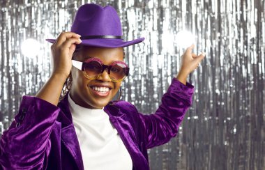 Mor parti kıyafetleri içindeki neşeli, şık, koyu tenli genç kadın disko düzenliyor. Parti gözlüklü, kadife ceketli ve arkasında parlak gümüş arka planda parmağını gösteren şapkalı bir kadın..