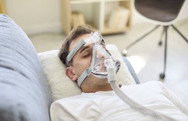 Aspiratörlü, oksijen maskeli bir adam yatakta dinleniyor. Hasta tedavi sırasında solunum desteği alıyor. Temiz kurulum, evde hastane sağlık hizmetleri olduğunu gösteriyor. Sakin tedavi konsepti.
