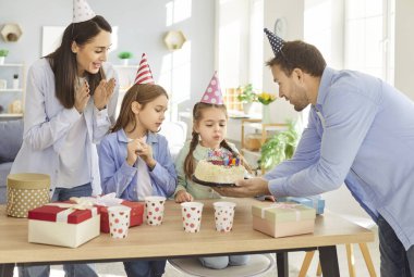Çocuklar partinin tadını çıkarırken mutlu aile evde doğum günü kutluyor. Genç kız pastaya mum üflüyor, neşeli aile kutlamalarına eğlence ve heyecan katıyor..