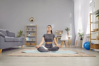 Genç yogi sakin, çekici kadın yoga yapıyor Ardha Padmasana egzersizi yapıyor, yarım Lotus pozu veriyor, mudra hareketleriyle meditasyon yapıyor, paspas üzerinde çalışıyor, oturma odasında spor kıyafetleri giyiyor.