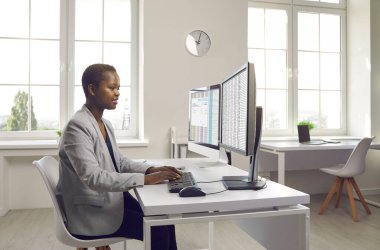 İş kadını ya da kiralık muhasebeci işte birden fazla bilgisayar kullanıyor. Takım elbiseli mutlu siyah kadın hesap tablosu ve fatura dosyalarıyla çalışıyor. Ofis masasında iki masaüstü bilgisayarıyla oturuyor.