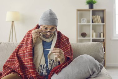 Grip ya da soğuk algınlığı belirtileri gösteren adam ateşini ölçer. Ateşi olan orta yaşlı hasta bir adam evdeki kanepede oturuyor ve şaşırmış yüz ifadesiyle termometreye bakıyor.