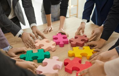 İş ekibi yapbozun parçalarını eşleştirmeye çalışıyor. Ahşap masanın etrafında duran ve renkli kırmızı, sarı, yeşil, pembe yapboz parçalarını birleştiren bir grup genç. Kesilmiş atış. Takım çalışması konsepti