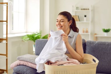 Kadın temiz çamaşır kokuyor. Kanepede çamaşır sepetiyle oturan, temiz, yıkanmış, beyaz gömlekli, taze kokan, doğal aromalı ve gülümseyen mutlu bir ev hanımı.