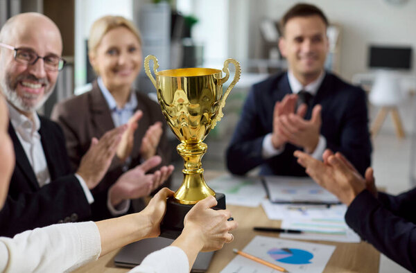 Деловая команда аплодирует награде и успеху. В офисе коллеги хлопают в честь празднования, когда кубок вручается победителю, демонстрирующему достижения и признание. Четкое представление о командной победе.