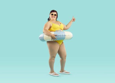Plaj lastiği yüzüklü mutlu ve şişman bir kadının portresi sarı mayo giymiş mavi bir stüdyonun arka planında başparmak işareti gösteriyor. Seyahat, tatil ve yaz yolculuğu konsepti.