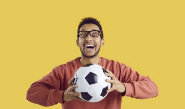Çılgın futbol taraftarı favori takımı destekliyor. Mutlu komik, heyecanlı, neşeli siyah adamın portresi koyu sarı arka planda izole bir şekilde duruyor, futbol topunu tutuyor, kameraya bakıyor ve gülümsüyor.