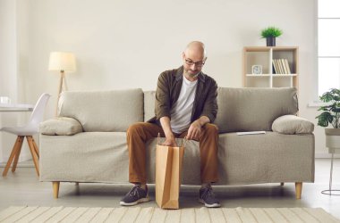 Müşteri kahverengi el işi kağıt teslimat çantasını açar. Orta yaşlı kel bir adam evdeki modern iç mekanda koltukta oturuyor ve internetten sipariş ettiği şeyleri açıyor. Çevrimiçi alışveriş ve hızlı teslimat kavramı