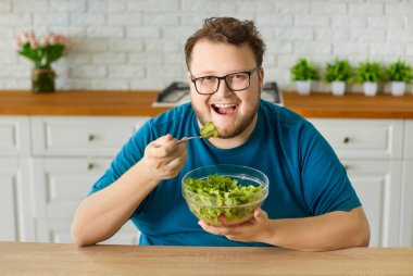 Gülümseyen adam diyette sağlıklı marul salatası yiyor. Parlak mutfakta büyük boy bir yetişkin cam kase ve çatal tutuyor, mutlu bir gülümsemeyle taze yeşilliklerin tadını çıkarıyor. Konsept: sağlıklı kilo verme motivasyonu.