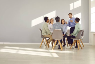 Profesyonel işletme koçu ve bir grup insanla takım danışmanı toplantısı. Mutlu adam, muhtelif erkek ve kadınlardan oluşan bir ekiple konuşuyor. Gri kopya uzay duvarıyla koltuklarda daireler çiziyorlar.