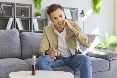 Alkol bağımlılığı olan bir adam stres altında sigara içiyor. Kanepede otururken, bir içkiye sarılıyor ve sorunu yansıtıyor, kötü sağlık, risk ve yükselen alışkanlık sinyalleri veriyor. Zararlı alkol alışkanlığı ve riski.