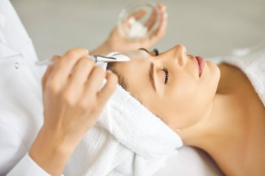 Kozmetik uzmanı, spa salonundaki genç, rahat, gülümseyen bir kadına nemlendirici maske takıyor. Güzellik kliniğindeki güzellik uzmanından güzellik prosedürü alan bir kız müşterinin portresini yakından çek