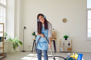 Evdeki oturma odasında elektrikli süpürgeyle yerleri temizleyen mutlu genç bir kadın. Ev hanımı kulaklıklı, evi süpüren müzik dinliyor. Temizlik ve temizlik hizmeti kavramı.