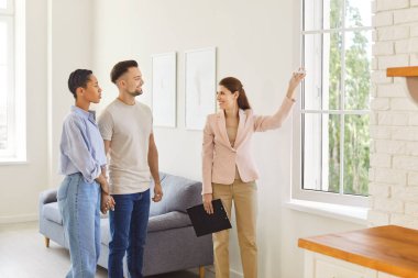 Gülümseyen kadın ajan, boş yeni dairelerde güneşli oturma odası ve pencere manzarasını gösteriyor. Emlakçı müşterilere yeni evi gösteriyor. Emlak, taşınma ve kira kavramı.
