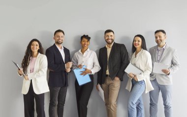 Gülümseyen bir grup iş adamı. Çeşitli bir şirket çalışanı gri bir duvarın yanında duruyor, elinde belgelerle, modern ofis kıyafetleri içinde. Tema takım çalışması, çeşitlilik, kurumsal başarı.