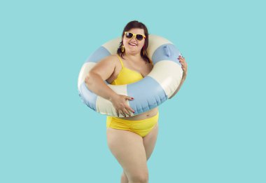 Plaj lastiği yüzüklü mutlu bir kadının portresi sarı mayo giymiş mavi bir stüdyonun arka planında izole edilmiş kameraya bakıyor. Seyahat, tatil ve yaz yolculuğu konsepti.