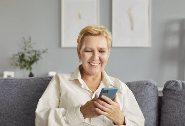 Evde boş vaktinin tadını çıkaran yaşlı bir kadın online eğlence ve mesajlaşma için akıllı telefon kullanıyor. Yurt içi rahatlama, internet bağlantısı ve huzurlu bir yaşam tarzı..