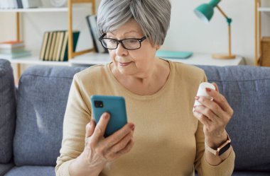 Yaşlı, gri saçlı, yaşlı bir kadının portresi elinde bir kavanoz tıbbi hapla koltukta oturuyor cep telefonunda uyuşturucu kullanma talimatlarını okuyor ya da internetten ilaç alıyor..