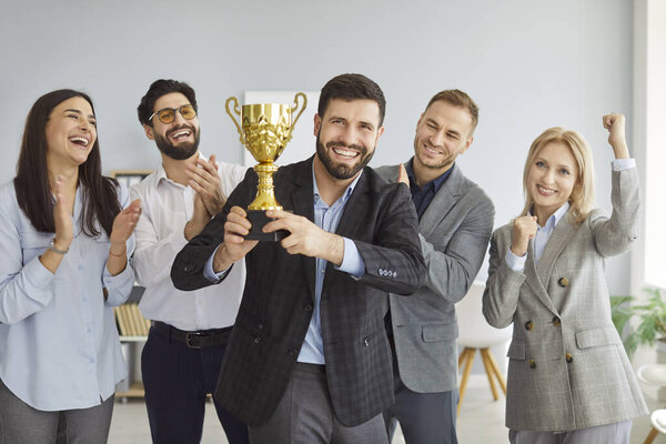 Портрет счастливого улыбающегося молодого человека-победителя, обладателя премии "Трофей" за успех в бизнесе, находящегося в должности, с командой коллег, аплодирующих лучшей награде сотрудников компании.