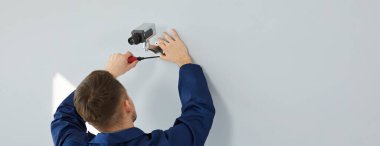 Back view of man using screwdriver while setting up new modern wall mounted security camera. Young male technician installing CCTV surveillance camera for theft deterrence inside residential building
