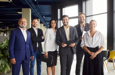 İş adamları modern ofisin açık alanlarında bir arada duruyorlar. Kendine güvenen genç ve yaşlı meslektaşlardan oluşan verimli bir ekip çalışması. Elinde kağıt kağıtlar olan panolar, gülümseyen insanlar.