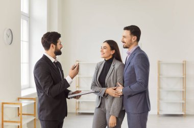 Dost canlısı emlakçı gülümseyen genç çifte modern bir daire ya da ofis alanı sunuyor. Emlakçı emlak seçeneklerini tartışıyor ve danışma sırasında potansiyel alıcılarla ayrıntıları satın alıyor..