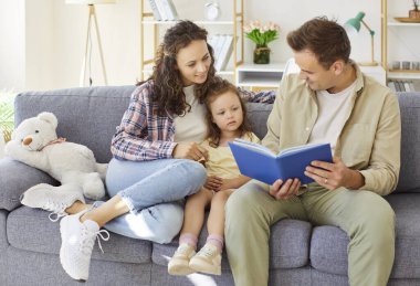 Mutlu ebeveynler ve küçük kız çocuk birlikte kitap okuyorlar. Evdeki rahat bir kanepede gülümserler, bir hikayeyi paylaşırlar ve yumuşak bir ayıya sarılırlar. Aile bağını, çocukluğu ve boş zamanlarını kutluyor.