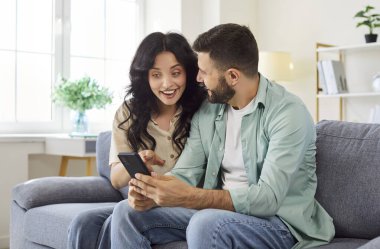 Mutlu çift iyi haberi kutlamak için akıllı telefonu kullanıyor. Evdeki rahat koltukta oturan kaygısız genç insanlar, mutlu bir şekilde telefon ekranına bakan ve pozitif mesajdan sürpriz yapan heyecanlı bir kadın.