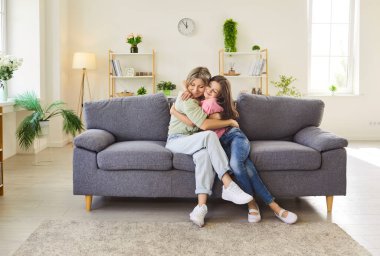 Anne ve kızı sevgiyle sarılıyorlar. Evdeki kanepeye yakından bakınca aile bağı, gülümseme ve oturma odasındaki ilişki görülüyor. Anne hamileyken sarılıyor. Sevgi, ilgi ve anneler günü.