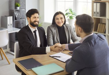 Ajan ya da mali danışman genç çiftle el sıkışıyor, anlaşma yapıyor, resmi bir toplantıda sözleşme imzalıyor. Banka kredisi ve kiracılık, satın alma ve satma hizmetleri, sigorta kavramı