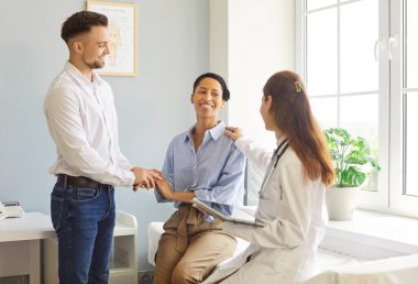 Gülümseyen bir aile doktoru muayene sırasında klinikteki kanepede oturan mutlu çift hastalarına destek oluyor. Kadın ve erkek, doktorla konuşuyor, tedavi için tavsiyeler alıyor..