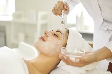 Kozmetik uzmanı, güzellik salonunda masaj fırçası kullanan bir müşteriye nemlendirici yüz maskesi takıyor. Uzman bakımı ve şımartılması, müşterinin cilt sağlığına titiz bir ilgi gösterilmesi,.