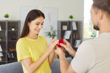 İçinde nişan yüzüğü olan küçük kırmızı bir yüzük taşıyan genç bir adam, güzel, sevgili kız arkadaşına evlenme teklif ediyor, ona evlenme teklif ediyor, gülümseyerek, evde romantik bir anın tadını çıkararak. Aşk, evlilik kavramı