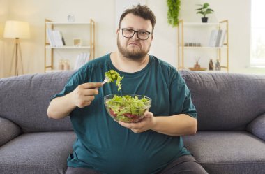 Evdeki kanepede salata yiyen mutsuz şişman şişman adam. Elinde bir kase yeşillik var, diyet konusunda isteksiz ve evde daha sağlıklı oturuyor. Sağlıklı beslenme, yemek, yaşam tarzı ve alışkanlıklar değişir..