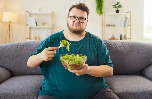 Evdeki kanepede salata yiyen mutsuz şişman şişman adam. Elinde bir kase yeşillik var, diyet konusunda isteksiz ve evde daha sağlıklı oturuyor. Sağlıklı beslenme, yemek, yaşam tarzı ve alışkanlıklar değişir..