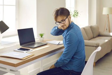 Oturarak yaşayan bir adam dizüstü bilgisayarda çalışırken boyun ağrısı hisseder. Yorgun bir şirket çalışanı dizüstü bilgisayarla masada oturuyor, el ele tutuşuyor, kaslarını rahatlatmak için masaj yapıyor.