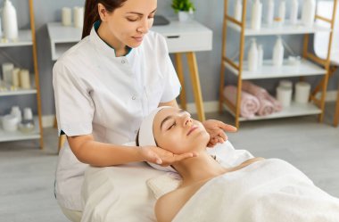 Estetisyen kozmetik uzmanı kadın müşterilerine spa salonunda yüz masajı yapar, cilt bakımı, rahatlama ve sağlığın önemini vurgular. Klinik stüdyosunda güzellik prosedürünün tadını çıkaran kişi.