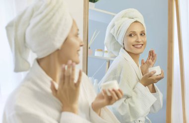 Mutlu sarışın kadın yüz kremi sürüyor, günlük yüz bakımı ve kaplıca güzellik rutininin tadını çıkarıyor. Bornoz ve havlu giymiş genç bir bayan banyo aynasında dikilip yansımaya ve gülümsemeye bakıyor.