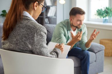 Hasta psikologla terapi seanslarındaki problemler hakkında konuşuyor, terapist modern ofiste dinleyip notlar yazıyor. Dürüst ve endişeli bir şekilde konuşan bir adam.