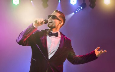 Sahnede mikrofonlu şarkıcı şovmen. Parlak spot ışıkları ve duman çerçevesi kadife bir takım elbise ve kendine güvenen bir adam şarkı söylerken papyon. Canlı eğlence konsepti ilham verici..