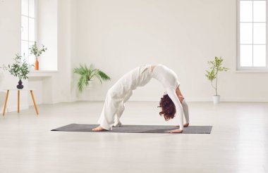 Yogi beautiful young woman working out in fitness club, home doing yoga pilates exercise. Bridge pose on mat, enjoying comfortable modern home interior design, backbend back, wearing white sportswear