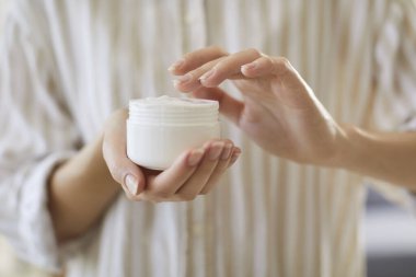 Kadın ellerinin yüz kremi dolu beyaz kavanozu tuttuğu, pürüzsüz, bakımlı cilt için organik cilt ürünleri uyguladığı yakın plan. Günlük kişisel bakım ritüeli, beslenme ve susuzluk.